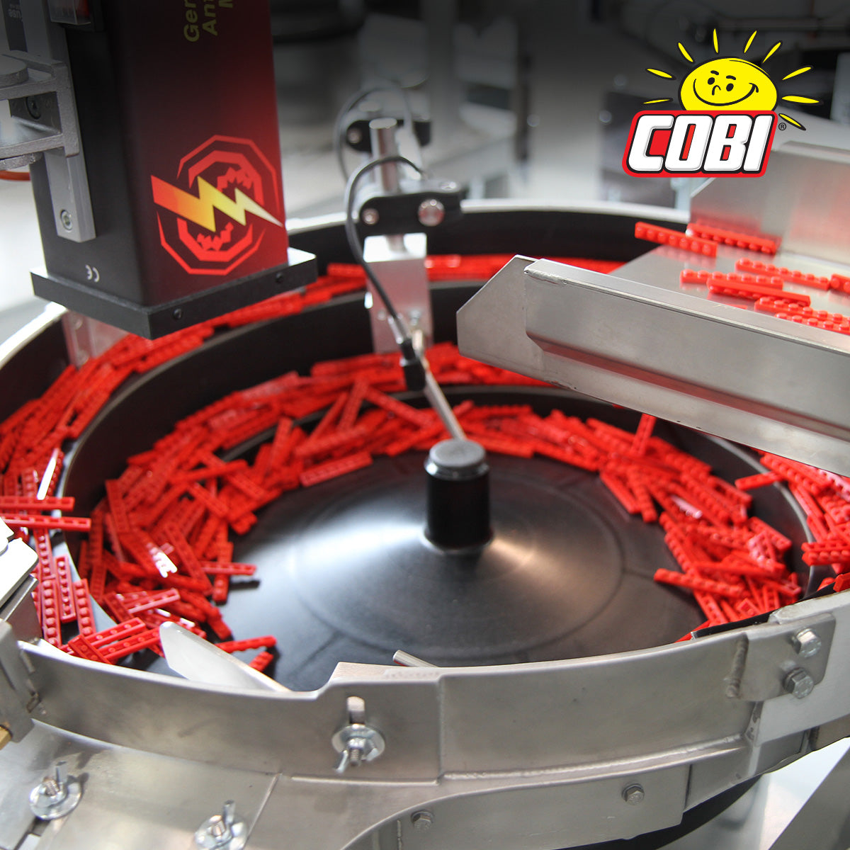 A close-up of a circular conveyor or sorting machine filled with many bright red plastic building blocks. The machine is made of silver metal with a black central hub and a red-and-black branded roller unit on the left.